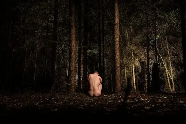 A nude figure in a protective fetal position at night, illuminated by a spotlight in a dark forest; a photographic metaphor for being overwhelmed by emotion.