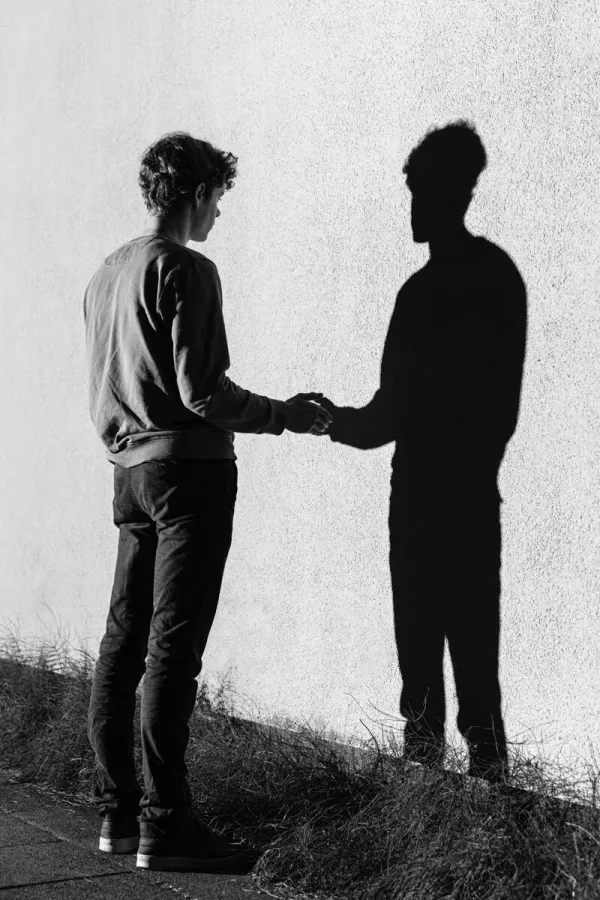 A black and white photograph of a man reaching out to shake hands with his own shadow on a textured wall.
